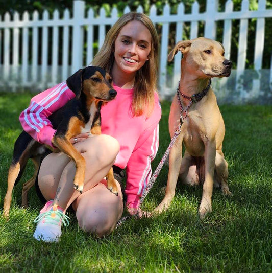 A smiling woman in a pink sweatshirt kneeling on grass with two dogs, one sitting on her lap and the other sitting beside her, in front of a white picket fence.